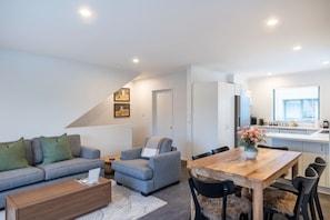 Ein modernes Wohnzimmer mit Sofa, Sessel, Couchtisch und Esstisch. Es gibt eine Küchenzeile mit Fenster und Kühlschrank.