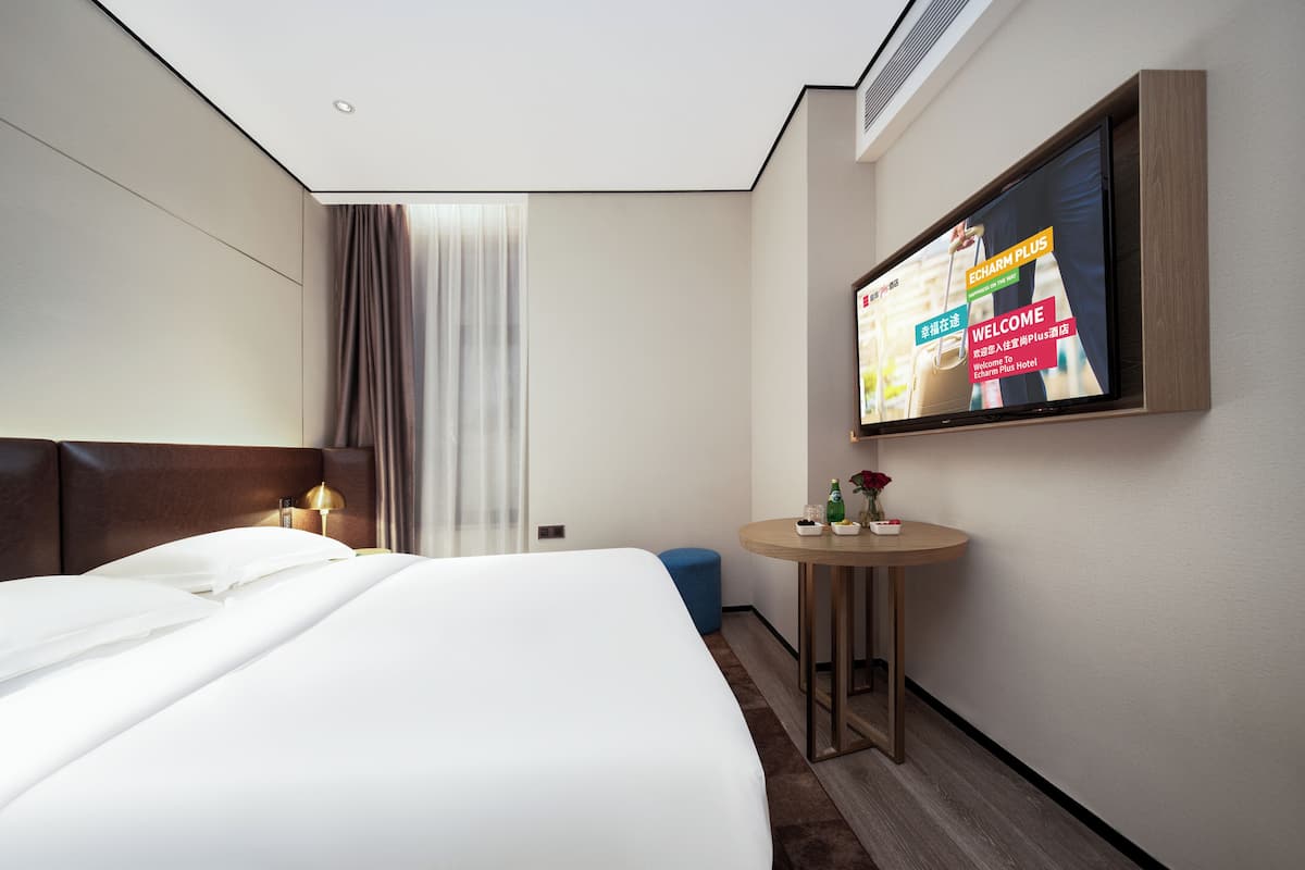A hotel room with a bed, a TV on the wall, a small round table, and a window with curtains.