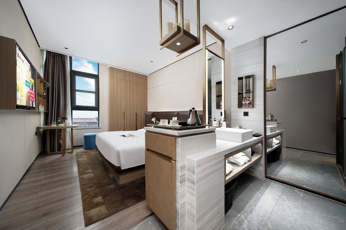 A modern hotel room with a bed, a bathroom with a mirror, and a window with curtains.