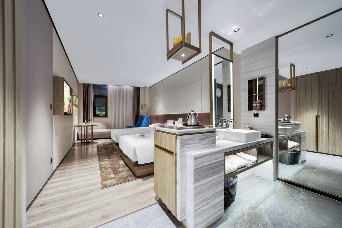 A modern hotel room with a large bed, a sitting area, a bathroom with a large mirror, and a small table with a lamp.