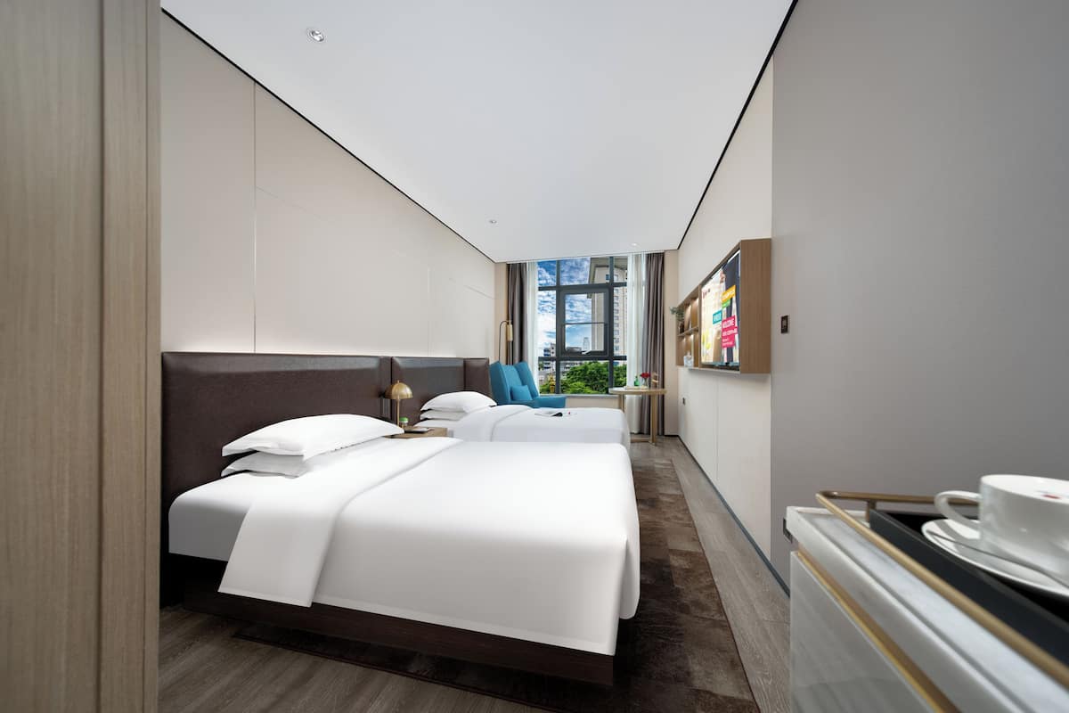 A modern hotel room with two beds, a large window, a small table with a cup, and a wall-mounted TV.