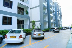 A multi-story residential building with parking spaces, a white car, and a silver car parked in front. - Cozy 2bed Master-ensuite apt in Nairobi Near the Airport and SGR Train Station (Syokimau)