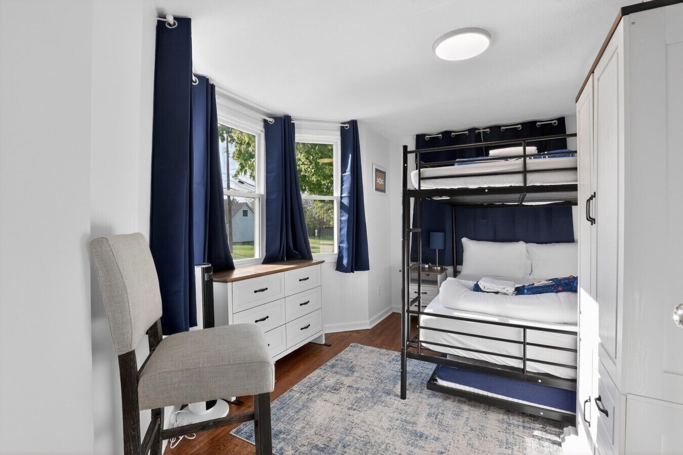 Habitación pequeña con literas, un escritorio con cajones, una silla y una ventana con cortinas.