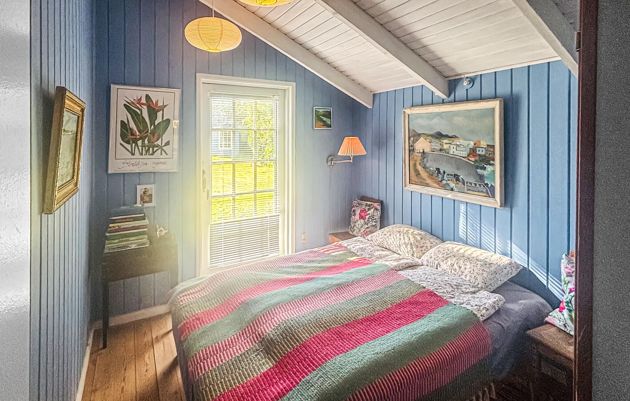 Ett sovrum med en säng, ett fönster med persienner, en lampa och konst på väggen.