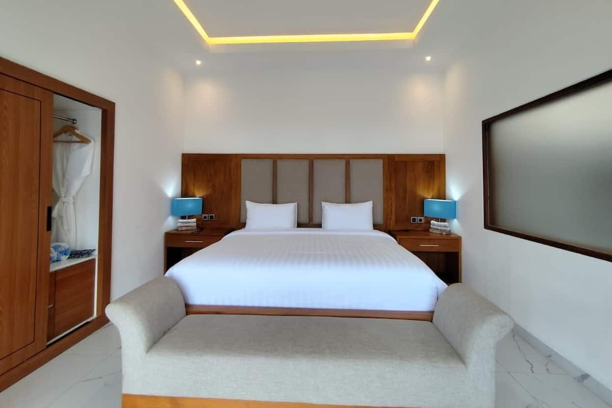 Una habitación de hotel moderna con una cama grande, cabecero de madera, mesitas de noche con lámparas, un armario y un televisor de gran tamaño.