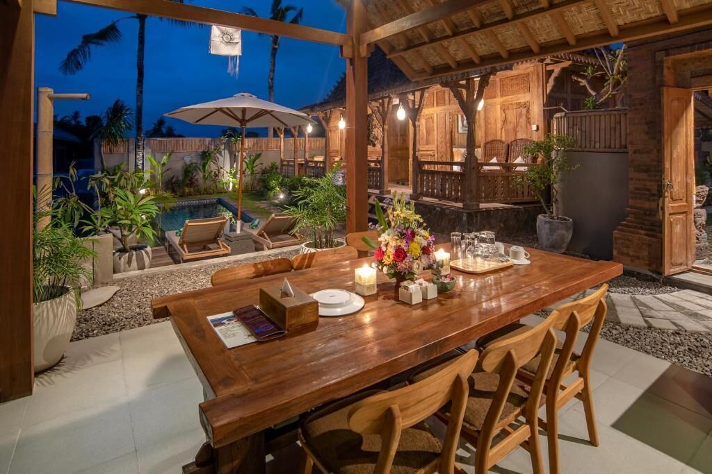 Uma área de refeições ao ar livre com uma mesa e cadeiras de madeira, e uma piscina ao fundo.