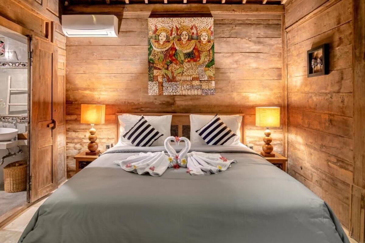 A wooden room with a bed, two bedside lamps, a wall tapestry, and a bathroom visible to the left.