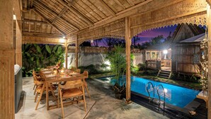 An outdoor dining area with a pool, wooden pergola, and tropical plants.