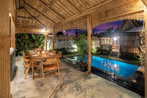 An outdoor dining area with a pool, wooden pergola, and tropical plants.