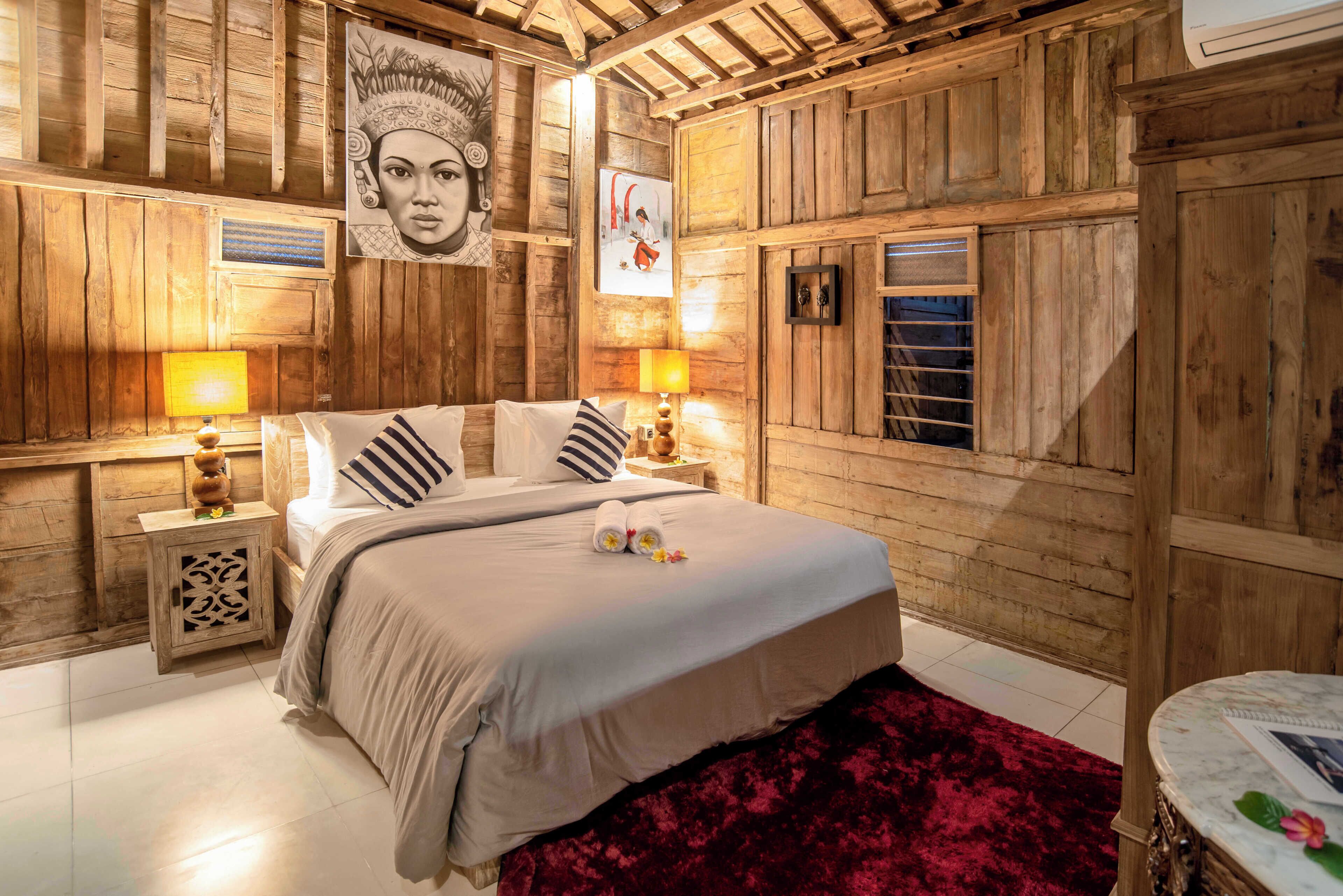 A wooden-paneled bedroom with a bed, two bedside lamps, a red rug, and artwork on the walls.
