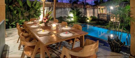 An outdoor dining area with a pool, wooden furniture, and tropical plants.