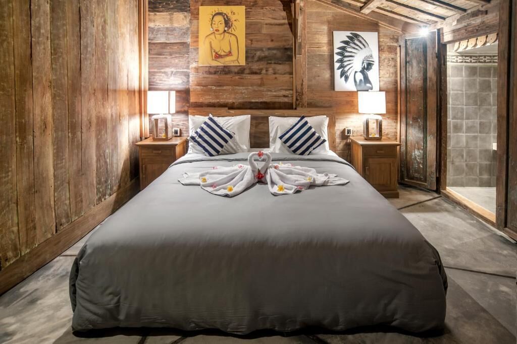 A large bed with a grey bedspread, two pillows, and a heart-shaped towel arrangement. There are wooden walls and a wooden ceiling.