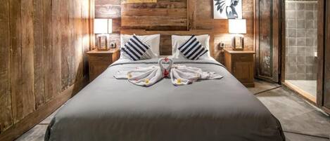 A large bed with a grey bedspread, two pillows, and a heart-shaped towel arrangement. There are wooden walls and a wooden ceiling.