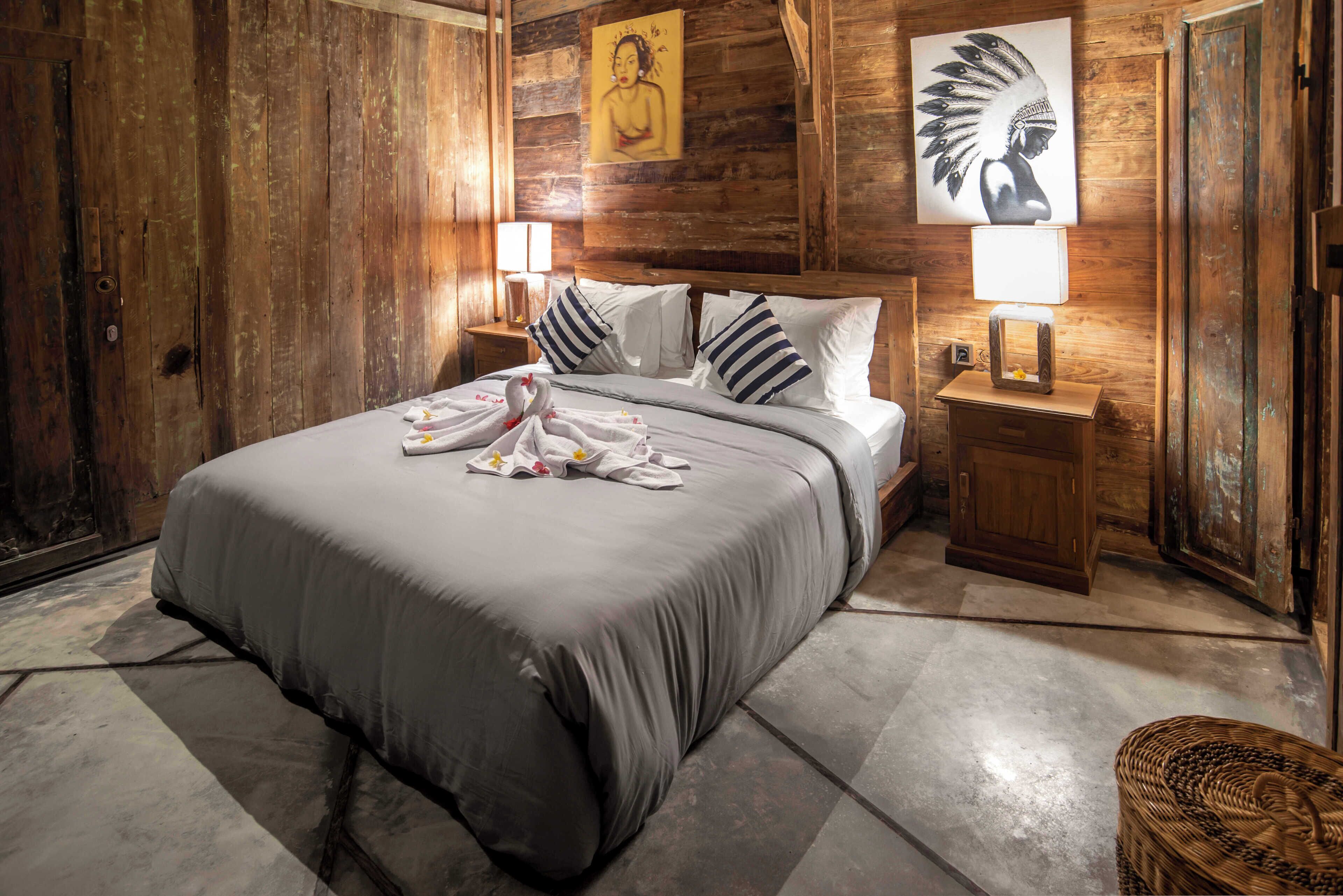 A room with a wooden wall, a bed with a grey bedspread, two nightstands with lamps, and a woven basket.