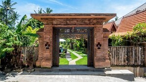 A brick entrance with a gate, a pathway leading to a building, and tropical vegetation.