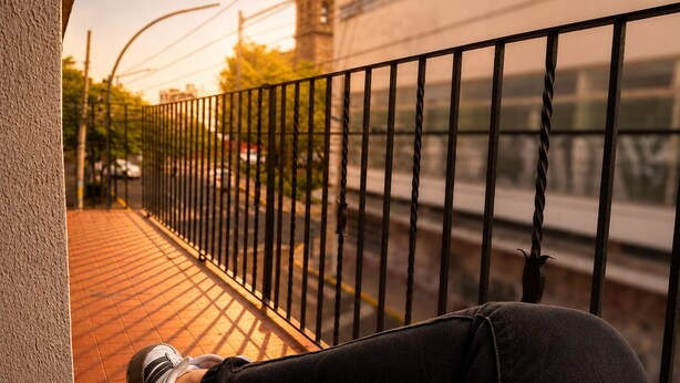Une personne assise sur un balcon doté d’une balustrade, tenant une tasse, et qui surplombe une rue où l’on aperçoit un train au loin.