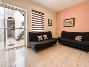 Ein modernes Wohnzimmer mit zwei dunklen Sofas, einem großen Fenster mit Jalousien, einem gerahmten Bild an der Wand und Blick auf eine Treppe, die zu einem zentralen Park führt.
