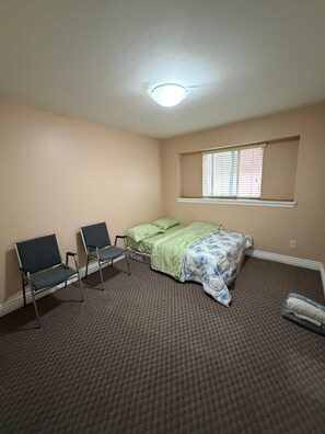 Quarto pequeno e simples, com uma cama, duas cadeiras, janela com persianas e chão coberto por carpete.