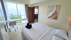 A hotel room with a bed, a desk, and a chair. There is a window with a view of greenery and a building. - Lux. 1BR Condo (55 SQM)  2 pools - Beach, Sea-views, Gym, Tours available (Lapu-Lapu City)