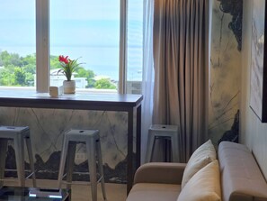 A room with a view of the sea, a sofa, a small table with a potted plant, and two stools.