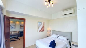 A hotel room with a bed, a nightstand, a lamp, an air conditioning unit, and a door leading to a sitting area with a sofa and a coffee table.