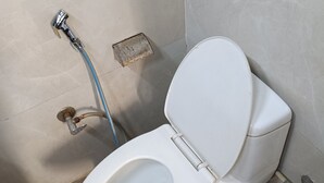 A white toilet with a closed seat, a visible flush handle, and a blue hose connected to a wall-mounted flush valve. - Hotel sudan inn kalka shimla higway  (Zirakpur)