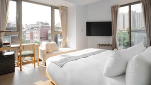 Una camera d'albergo con un letto, una scrivania, una sedia, una TV e ampie finestre con tende.