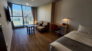 A hotel room with a bed, a desk with a chair, a sofa, a TV, and a large window with a view of buildings.