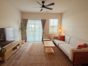 Uma sala de estar com televisão de tela plana, ventilador de teto, sofá, mesa de centro e uma janela grande com cortinas.