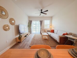 Un salon moderne doté d’un téléviseur à écran plat, d’un ventilateur de plafond, d’un canapé, d’une table à manger avec des chaises et d’une grande fenêtre avec des rideaux.