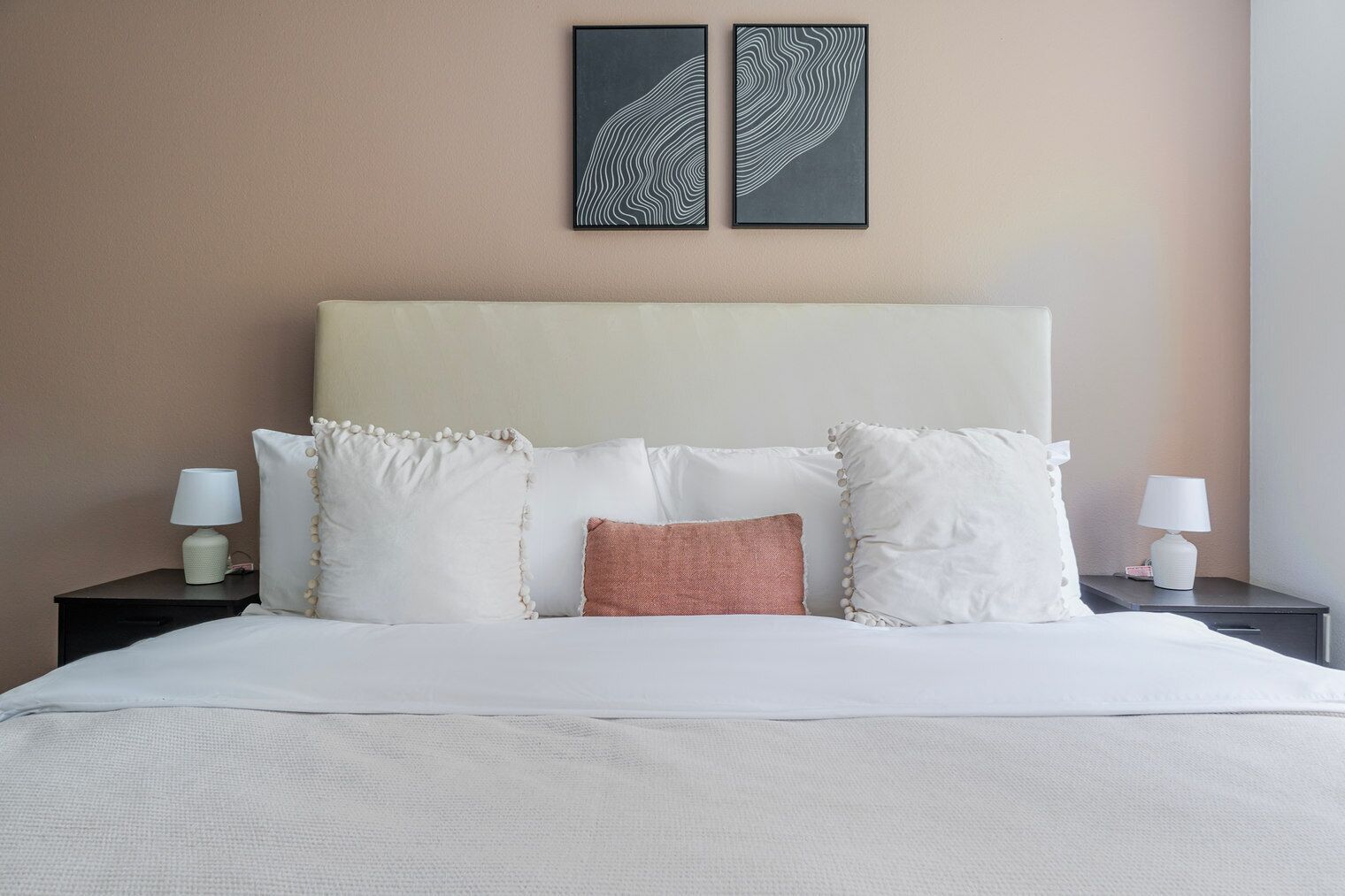 Cama bem arrumada com almofadas brancas e cor-de-rosa, cabeceira bege e duas pinturas abstratas penduradas na parede.