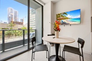 Ein modernes Zimmer mit rundem Tisch, zwei Stühlen, einem Fernsehgerät an der Wand und einem Balkon mit Blick auf die Stadt.