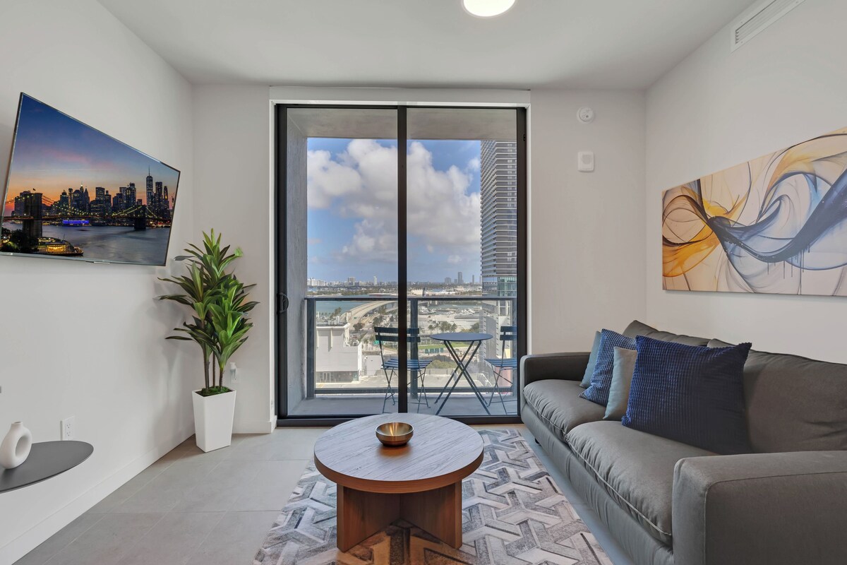 A modern living room with a sofa, a coffee table, a potted plant, a painting, and a large window with a city view.