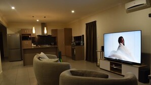 Een moderne woonkamer met een flatscreen televisie, twee beige fauteuils, een keukenhoek en een eettafel.