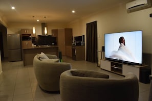 A modern living room with a flat-screen TV, two beige armchairs, a kitchen area, and a dining table. - The Duncan Retreat Bald Hills QLD (Bald Hills)
