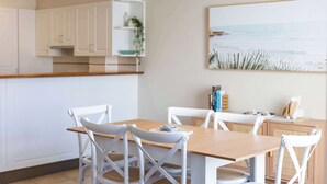 Un comedor con una mesa de madera y sillas blancas, una cocina con armarios blancos y un cuadro con temática de playa en la pared.