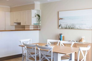 A dining area with a wooden table and white chairs, a kitchen with white cabinets, and a beach-themed painting on the wall. - Elanora- Oceanstays- Breakers Pippi Beach (Yamba)