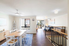 A spacious living area with a dining table, chairs, a blue patterned rug, a staircase with a railing, and a view to an outdoor patio. - Elanora- Oceanstays- Breakers Pippi Beach (Yamba)