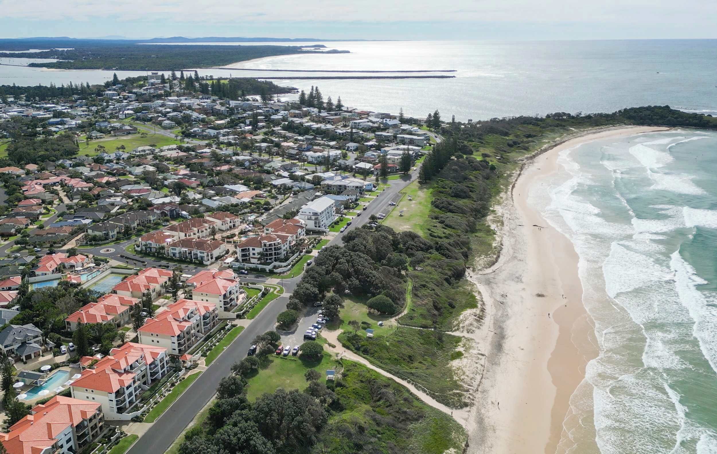 An aerial view of a coastal residential area with houses, a beach, and a bridge in the background.