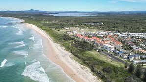 Vista aérea de una zona costera con playa, edificios residenciales y una cadena montañosa distante.