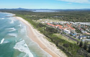 An aerial view of a coastal area with a beach, residential buildings, and a distant mountain range. - Elanora- Oceanstays- Breakers Pippi Beach (Yamba)