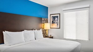 Une chambre d’hôtel avec un lit, une table de chevet, une lampe, un tableau encadré et une fenêtre avec des stores.