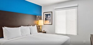 Et hotelværelse med en seng, et natbord, en lampe, et billede i ramme og et vindue med persienner.