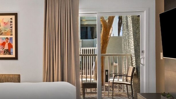 Une chambre d’hôtel avec un lit, une porte-fenêtre coulissante donnant sur un balcon, un tableau encadré au mur et un téléviseur à écran plat fixé au mur.