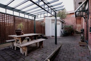 Un'area patio coperta con un tavolo e panche di legno, una recinzione bianca e un muro in mattoni.