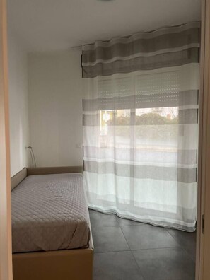 Un dormitorio con una cama, un ventanal con cortinas translúcidas y piso de baldosas.