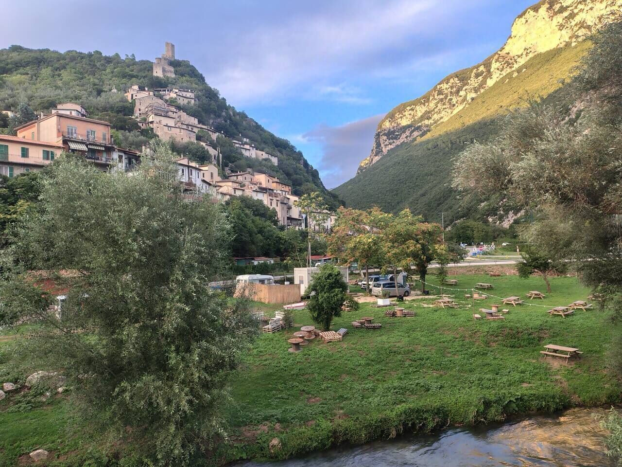 Un pueblo en una ladera con edificios, un río y mesas de picnic.