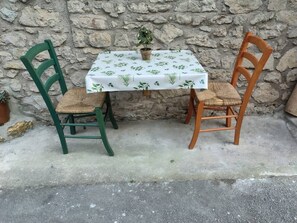 Une table recouverte d’une nappe blanche, avec deux chaises, une verte et une marron, devant un mur de pierre.