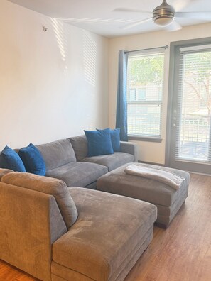 A living room with a grey sectional sofa, a small ottoman, and a ceiling fan. - Cozy 1-bedroom apartment near addison square (Dallas)