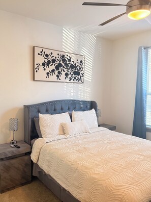 A bedroom with a tufted headboard, a bed with white bedding, a framed floral artwork, a bedside lamp, and a ceiling fan. - Cozy 1-bedroom apartment near addison square (Dallas)
